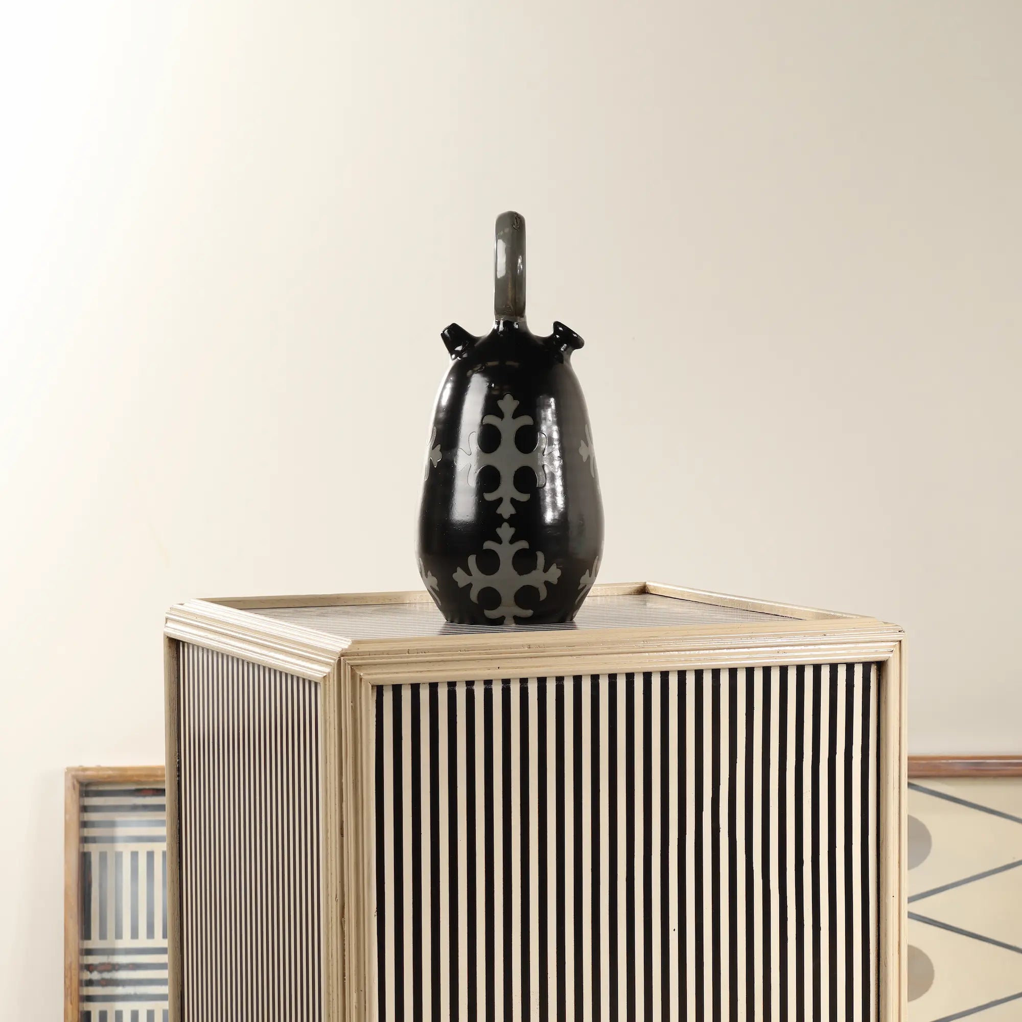 Botijo Ovum de cerámica negra colocado sobre un mueble decorativo de rayas en un ambiente interior.