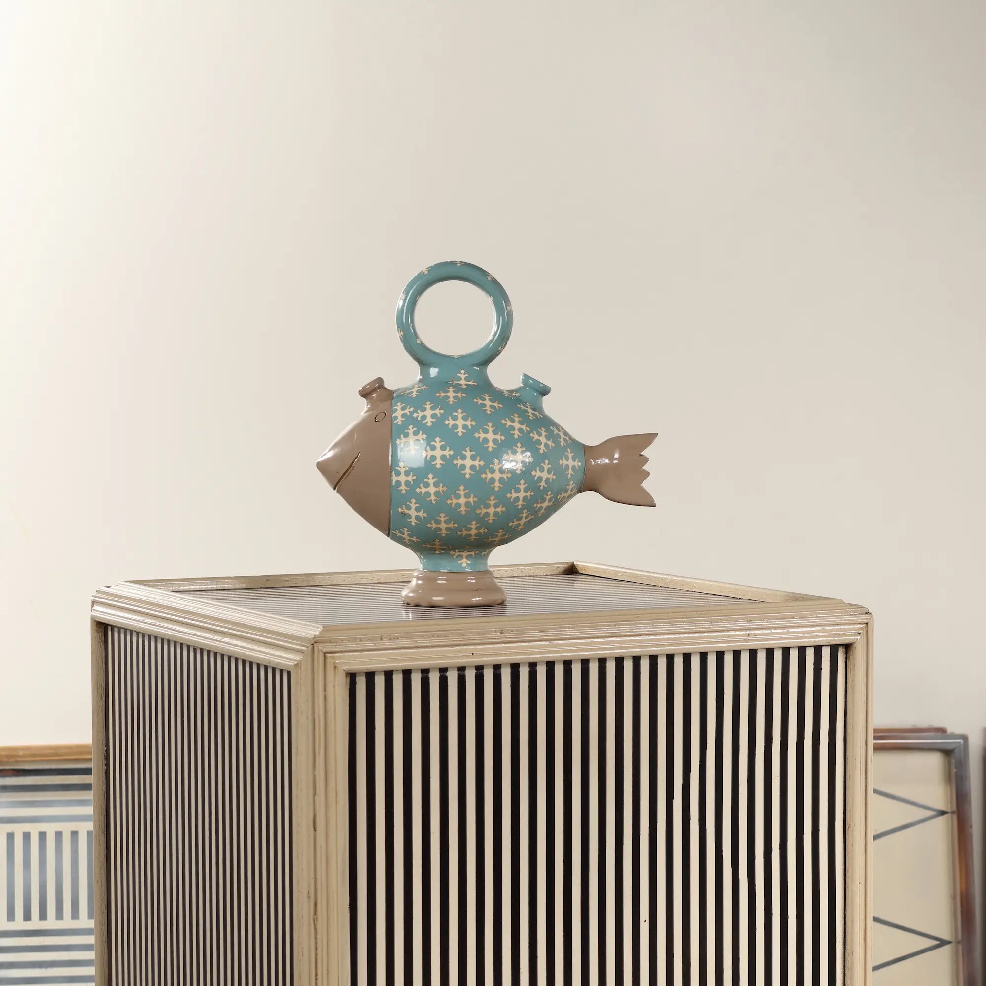 Botijo Poisson de cerámica colocado sobre un mueble decorativo de rayas, como pieza original para salones o recibidores.