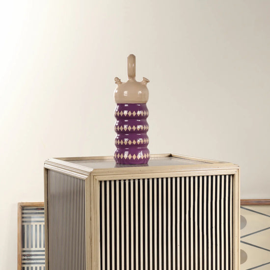 Botijo Rombos de cerámica morada colocado sobre un mueble decorativo de rayas en un ambiente interior.