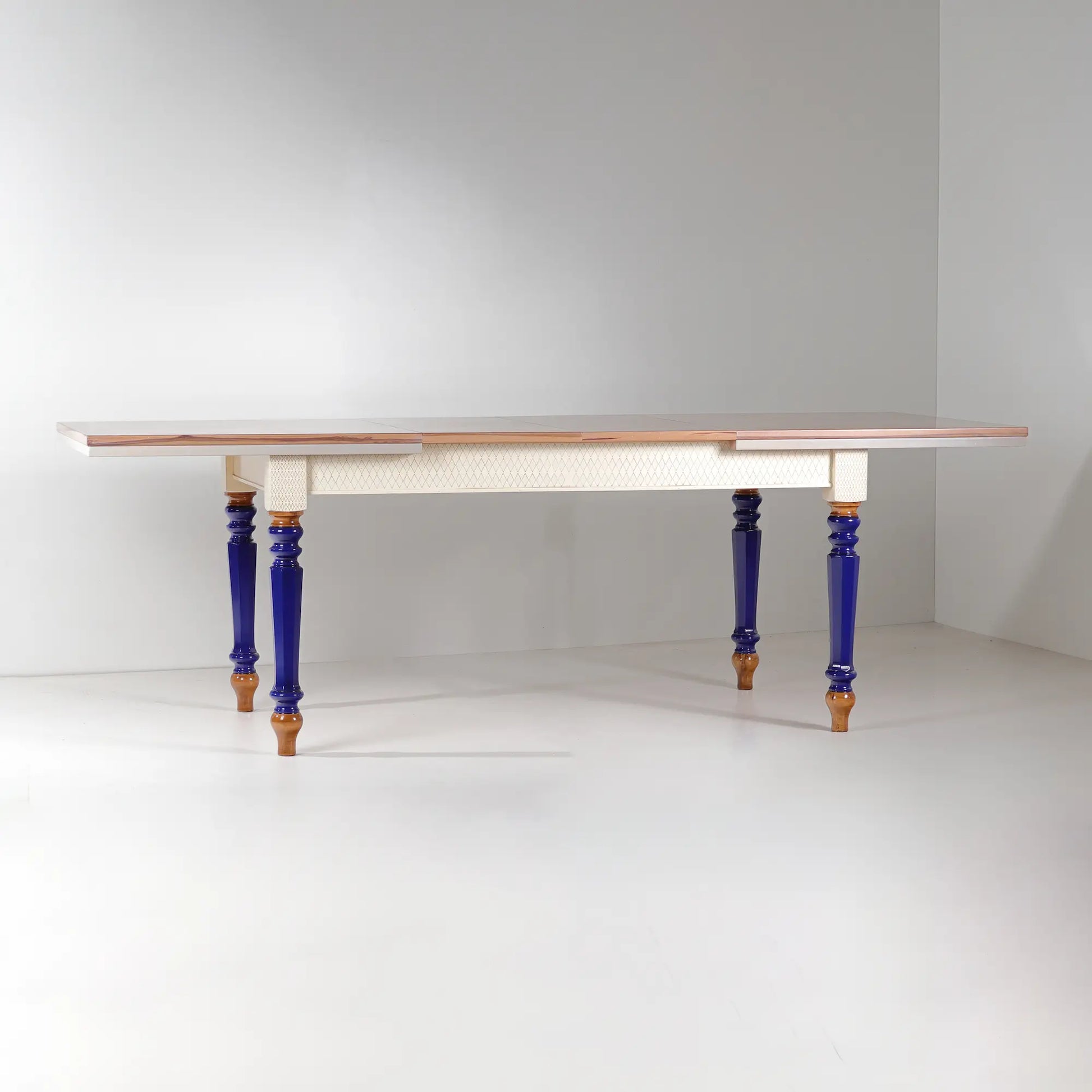 Mesa de comedor extensible Cascabella con sobre geométrico, faldón blanco y patas azul cobalto.