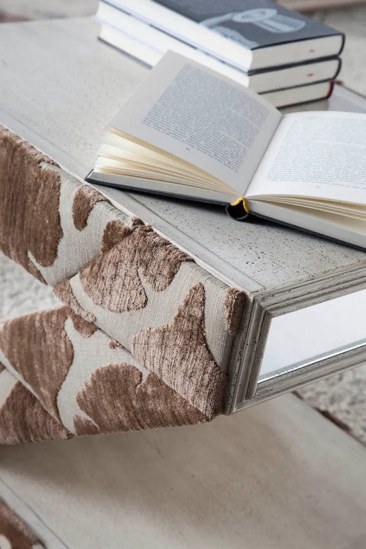 Detalle de la Mesita Z con acabado textil en tonos tierra, superficie envejecida y libro abierto sobre la mesa auxiliar de diseño.