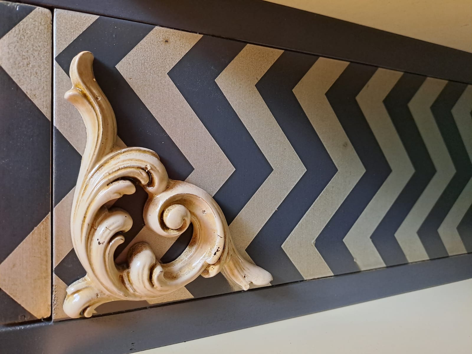 Detalle del Sinfonier Alice con aplique barroco en madera y patrón chevron en tonos oscuros.